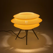 ORBIT Bone lit on a pale surface, three stacked knit-textured discs glowing warm above black tripod legs