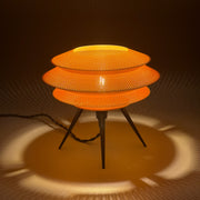 ORBIT Caramel lit on a pale surface, three stacked knit-textured discs glowing warm above black tripod legs